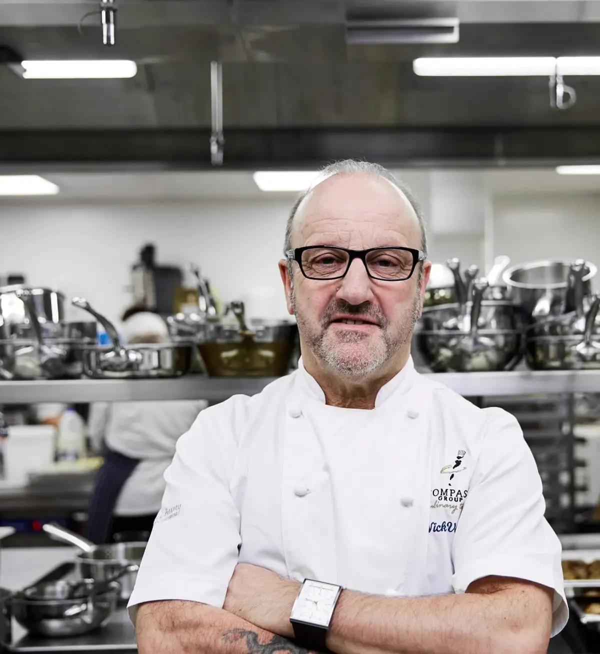 A chef in whites standing with arms folded in a commercial kitchen, racks of steel pans on the shelves behind