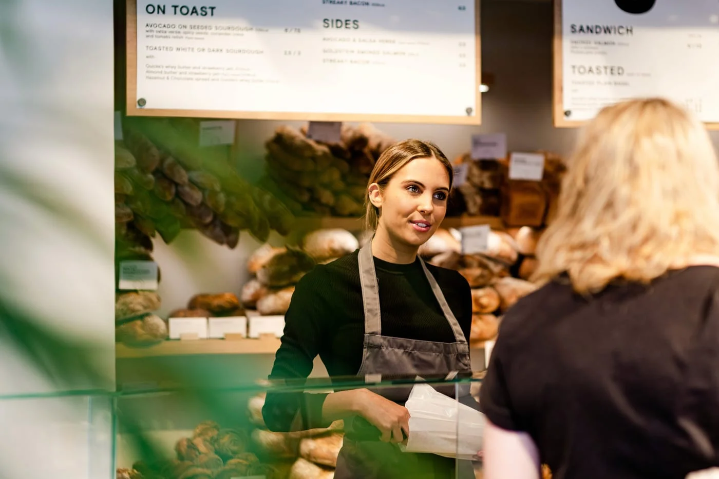 A bakery counter during service, loaves stacked on the wall behind and a menu board overhead