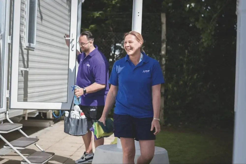 Two holiday park staff walking between caravans on a summer morning, cleaning kit in hand