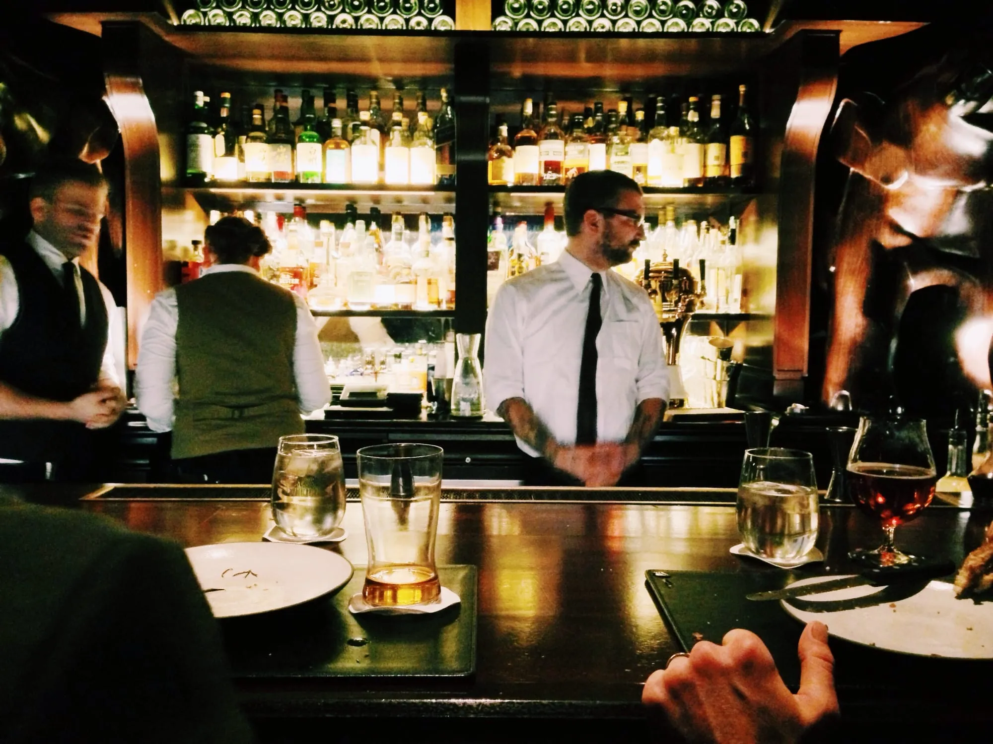 Bartenders at work behind a dimly lit bar lined with bottles, seen from a seat at the counter.