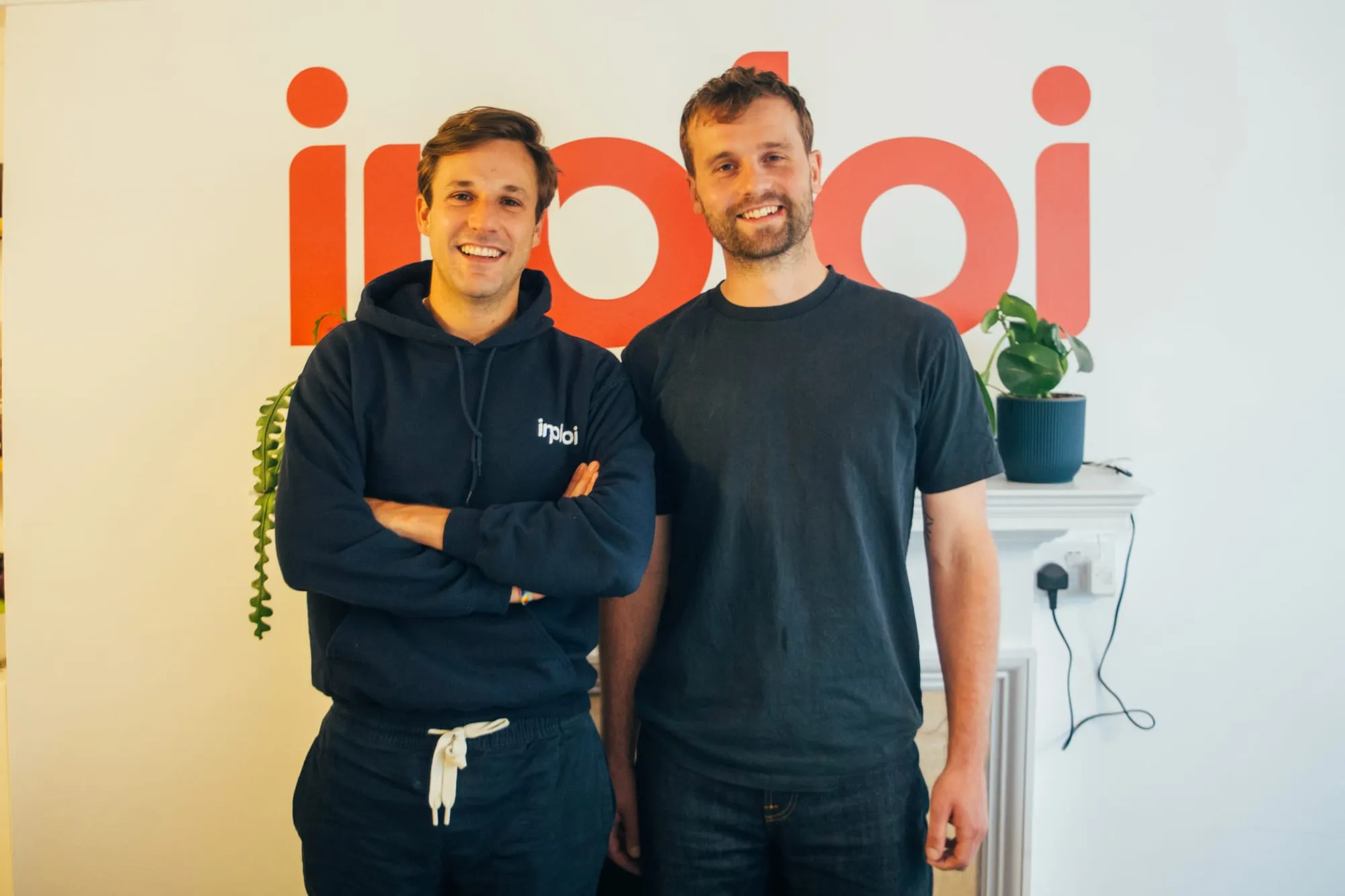Two people smiling in front of a large inploi logo on an office wall.