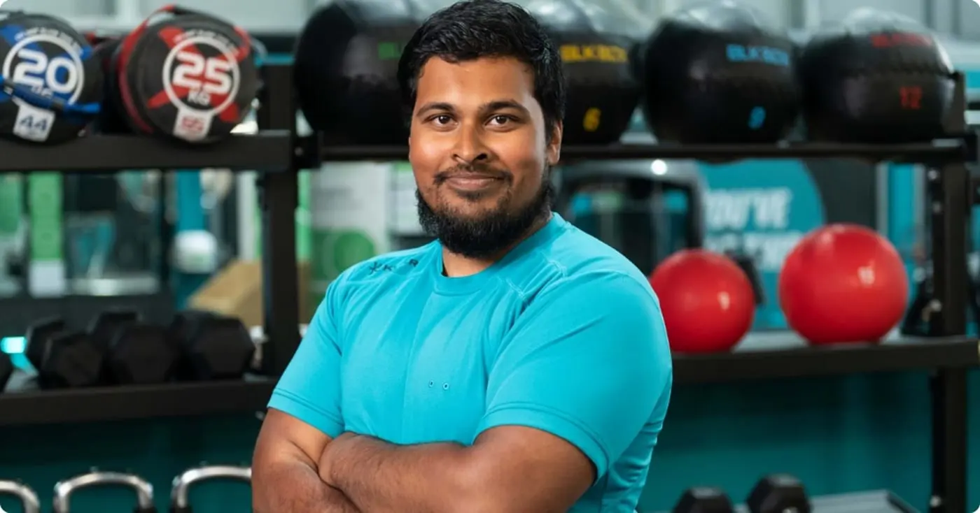 A person in a turquoise T-shirt standing with arms folded on a gym floor, medicine balls and dumbbells racked on the shelves behind