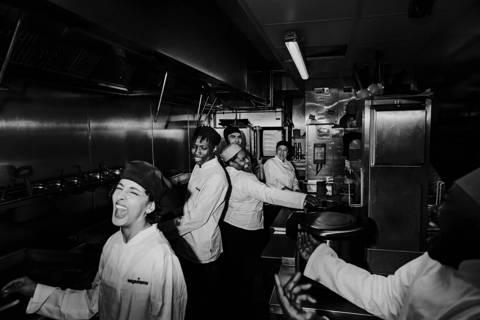 A restaurant kitchen brigade mid-service in black and white, dishes going out along the pass under the extraction hood